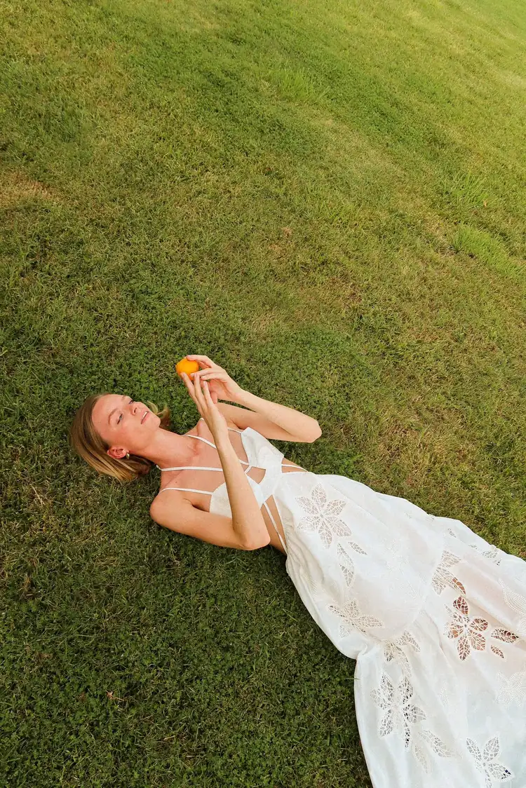A woman in a white embroidered dress lying on the grass, holding an orange